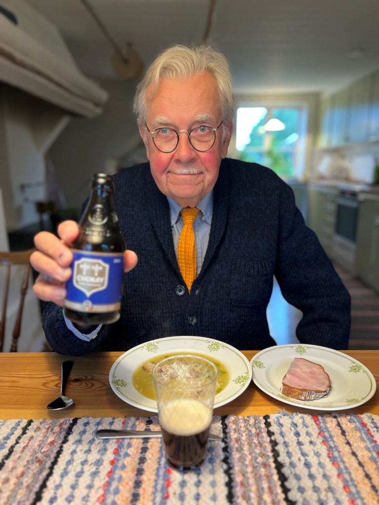 En äldre man sitter vid ett bord med en tallrik mat och en glas öl. Han håller upp en flaska öl och ler mot kameran. I bakgrunden syns ett kök med gröna skåp.