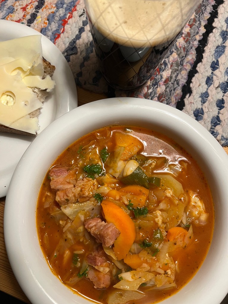 En skål med grönsaks- och köttsoppa garnished med färsk persilja, serverad med en skiva mörkt bröd med ost och en glas med mörk dryck, på en texturerad matta.