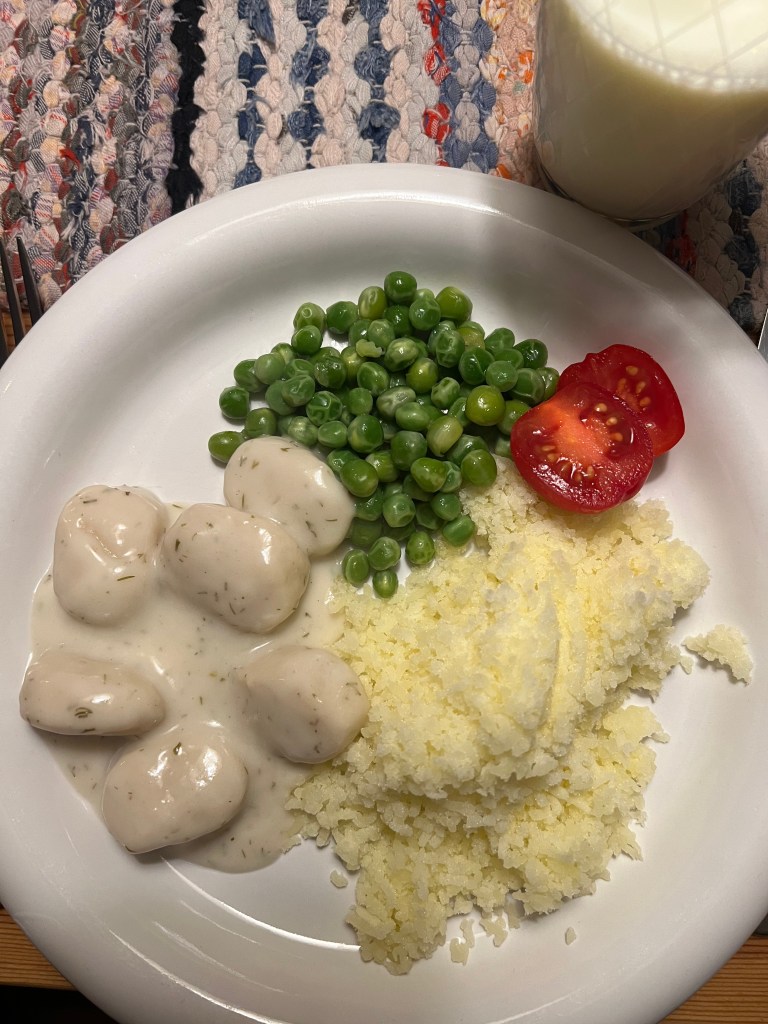 En tallrik med fiskbullar i sås, potatismos, ärtor och skivade tomater, serverad med ett glas mjölk i bakgrunden.