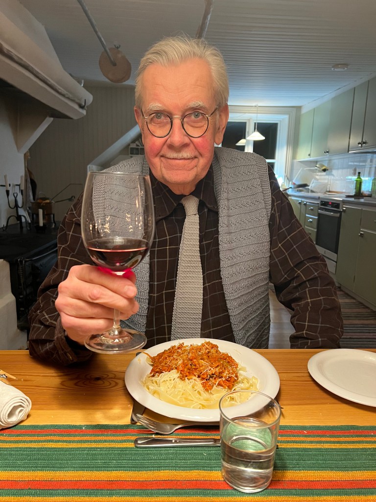 En man i glasögon och grått hår sitter vid ett bord med en tallrik spaghetti med sås framför sig, håller upp ett glas rödvin och ler.