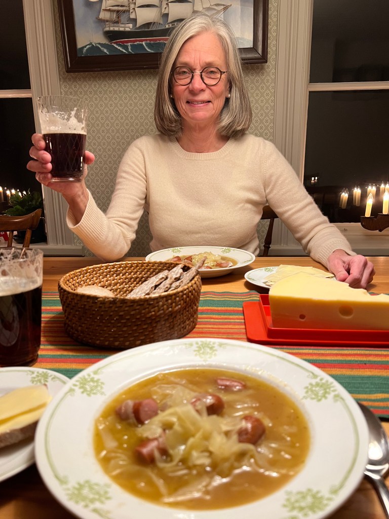 En kvinna som sitter vid ett bord med en skål sopa innehållande korv och kål, och höjer ett glas med dryck. På bordet finns också en brödkorg med knäcke, ost och ett fat med smör.