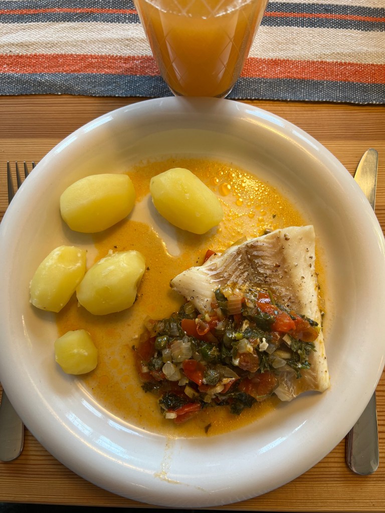 En tallrik med potatis, en bit fisk i sås och grönsaker, samt ett glas juice på en träytan.