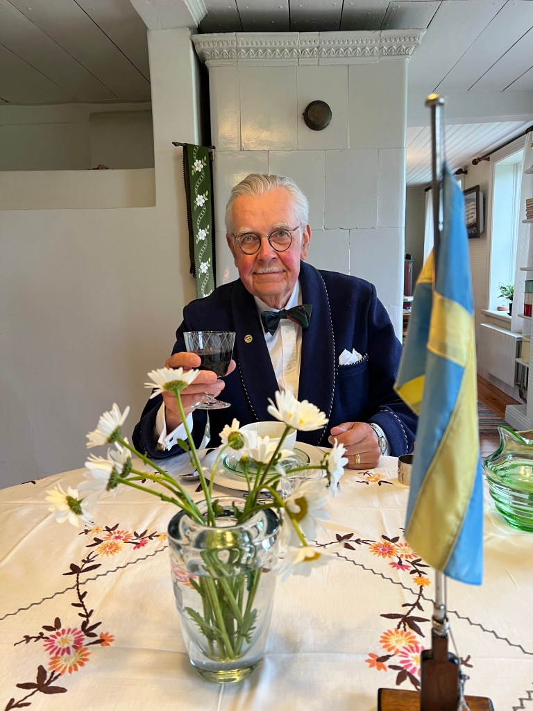 En man sitter vid ett bord med en duk dekorerad i blommor. Han håller upp ett glas och ser glad ut. På bordet finns en vas med blommor, en tallrik och en liten svensk flagga.
