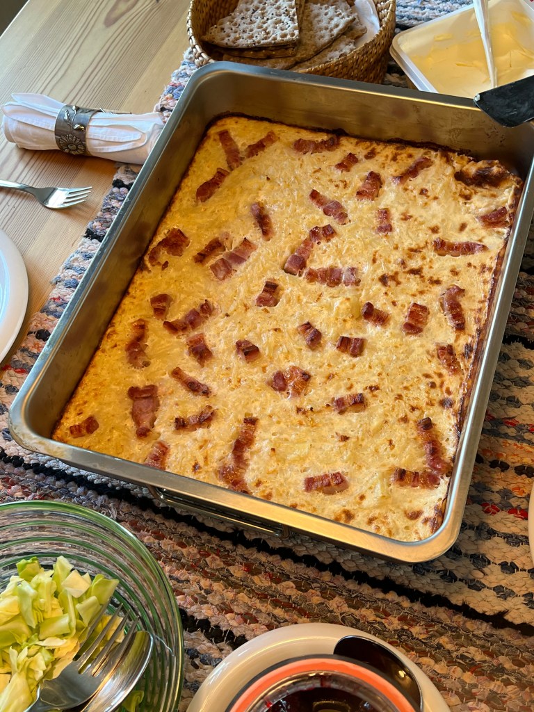 En ugnsrätt med en gyllene yta, toppad med skivor av bacon, sitter i en metallform på ett dukat bord. Vid sidan finns en skål med grönsallad, en tallrik och en burk med smör.