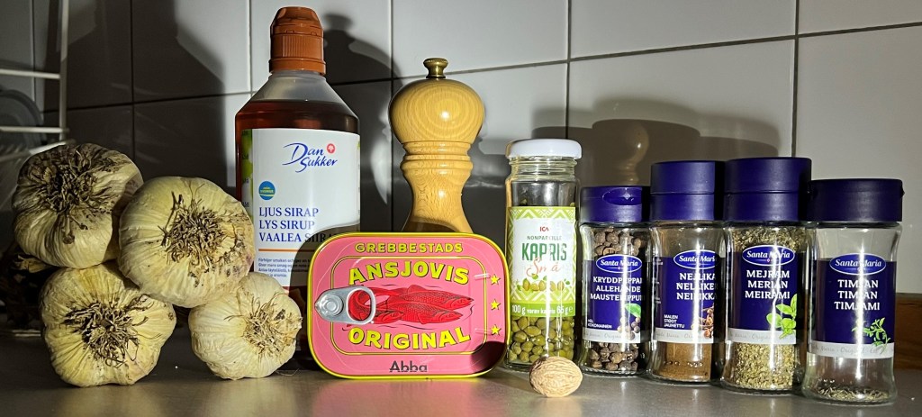 En köksbänk med vitlök, ansjovis i burk, kapris, kryddor som muskotnöt, svartpeppar, mejram, timjan, samt sirap på en grå yta.
