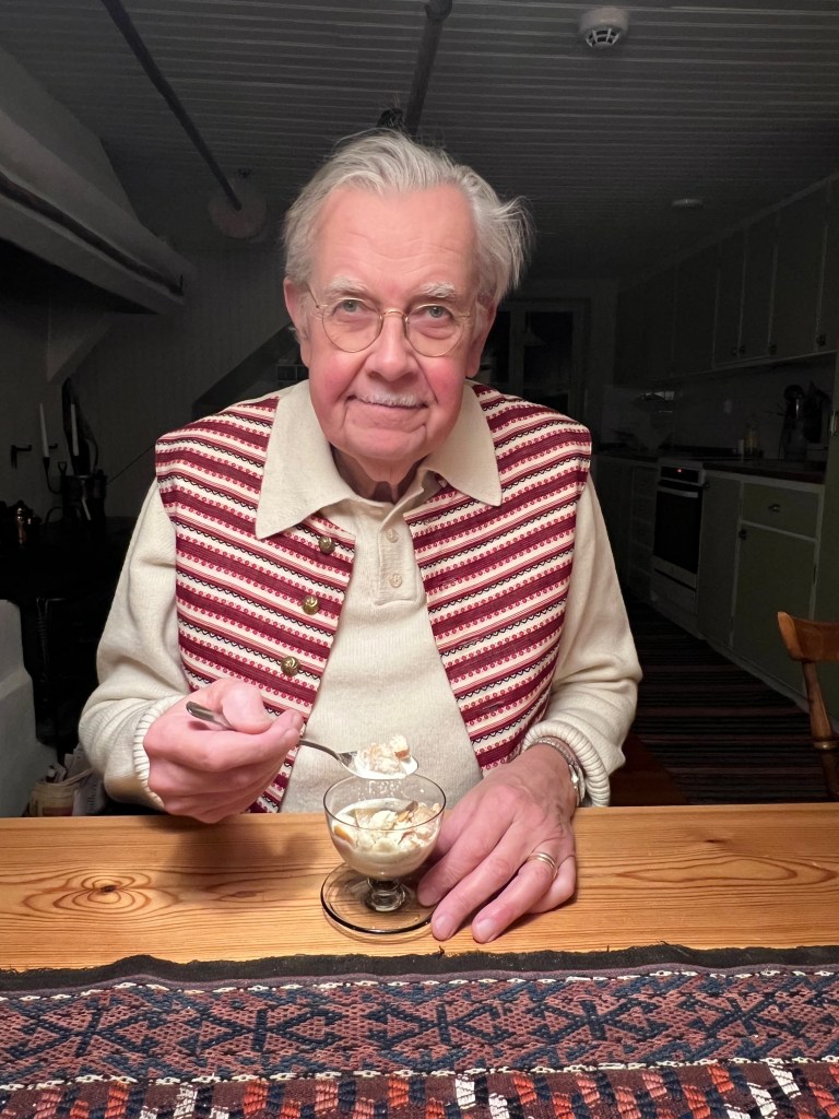 En äldre man sitter vid ett bord och äter glass ur en liten skål, iförd en randig väst och ser glad ut.