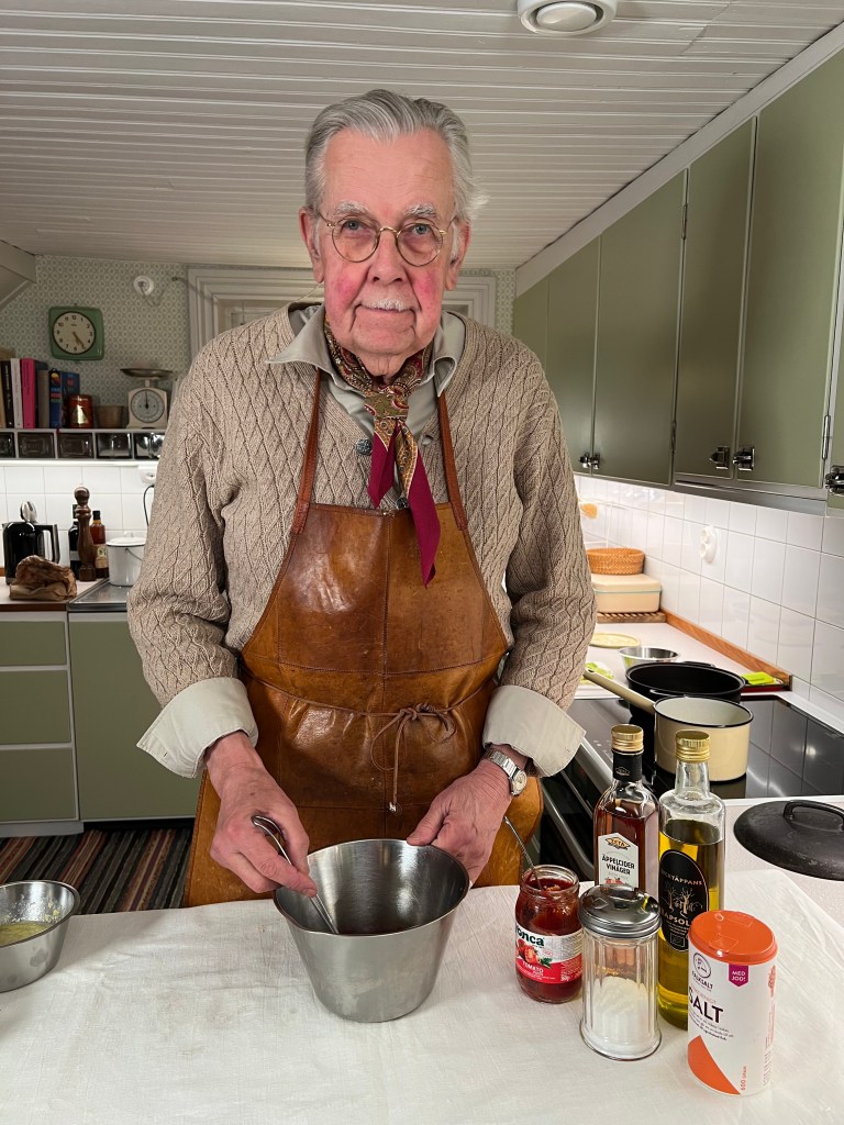 En äldre man i en köksmiljö, iförd en läderförkläde och glasögon, står vid ett bord och rör i en metallskål. Runt honom finns ingredienser såsom sylt, olja och salt.