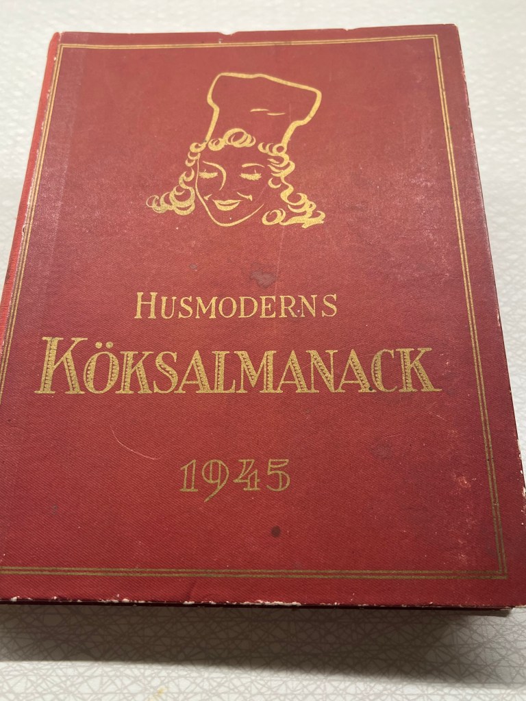 En gammal kokbok med titeln 'Husmoderens Kökssalmanack' från 1945, med en illustration av en kvinna i kockmössa på framsidan.
