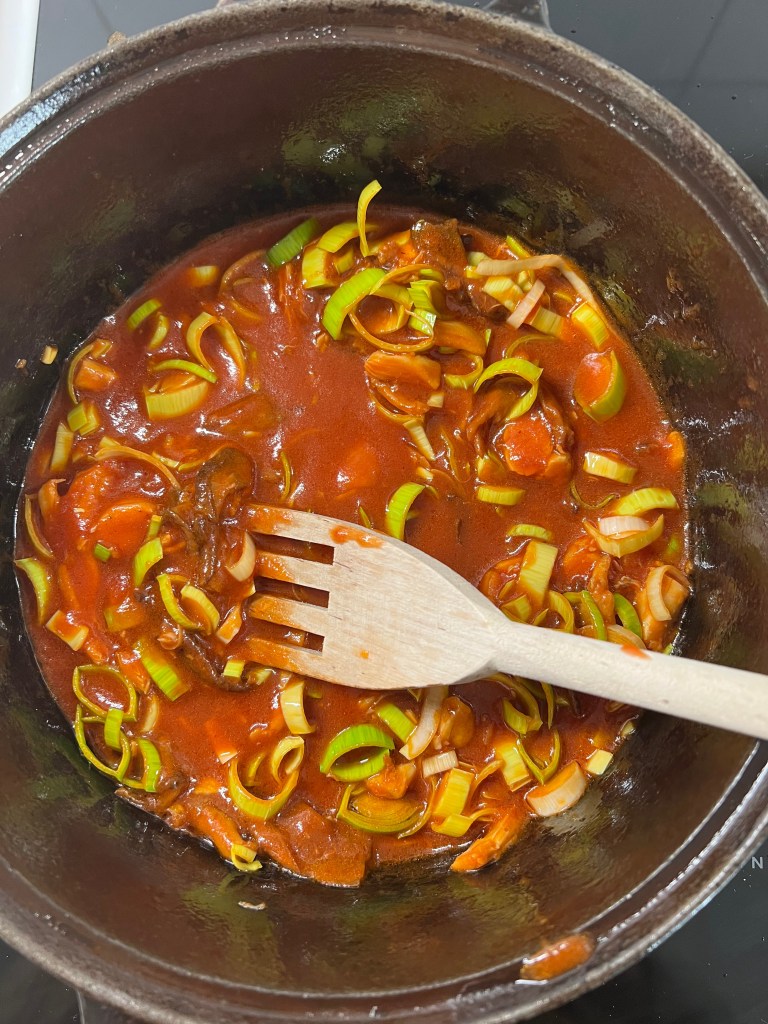 En kastrull med en tomatsås som innehåller skivade purjolökar och en träsked.