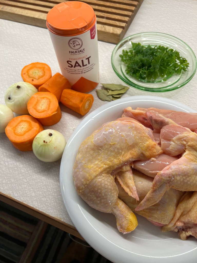 En platta med rå kycklingdelar, morötter, lök, en burk med fint salt och färsk koriander på ett bord.