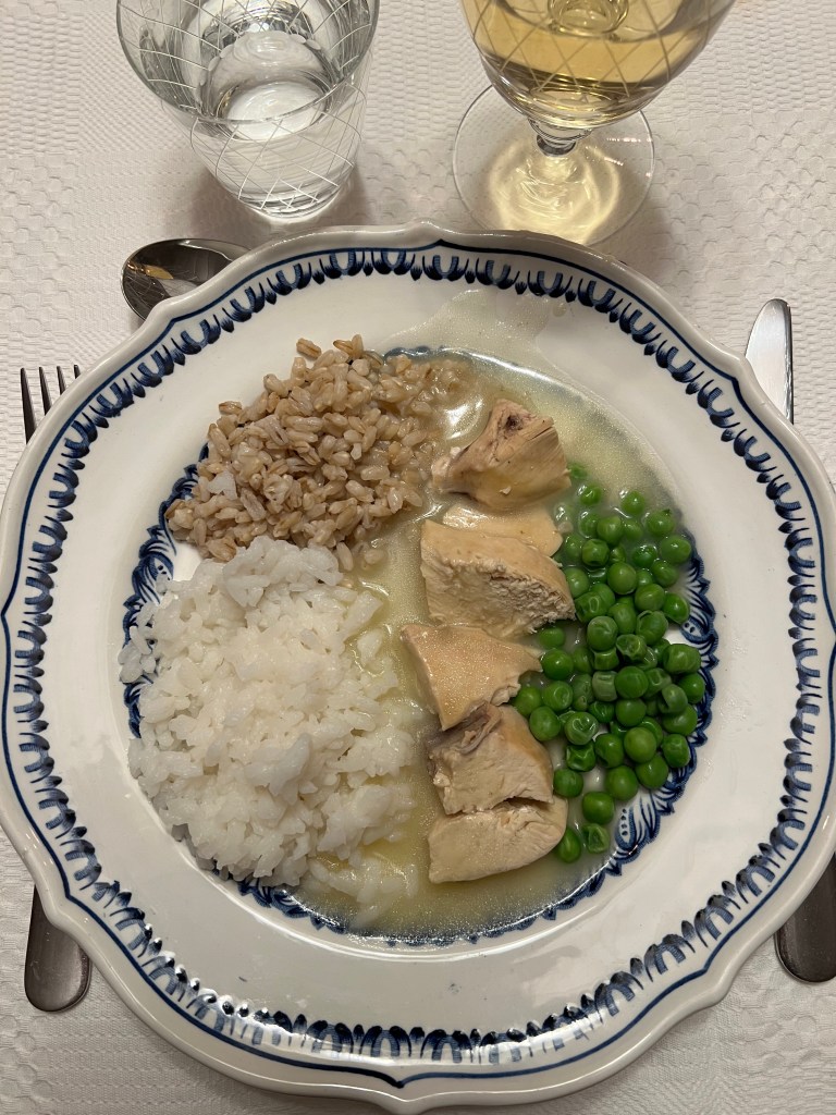 En tallrik med ris, korn, kycklingbitar och gröna ärtor, serverad med ett glas vitt vin och ett vattenglas.