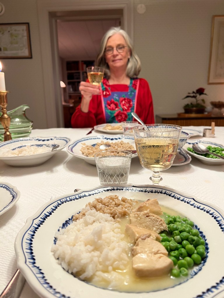 En kvinna med grått hår, klädd i en röd blus och blomsterförkläde, håller upp ett glas med dryck vid ett middagsbord. På tallriken framför henne finns ris, kyckling och gröna ärtor.