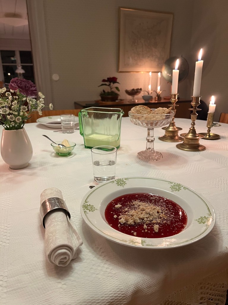Ett elegant dukat bord med en skål röd soppa toppad med smulor, ett glas vatten, en grön kanna och tända ljus i brassestakar.