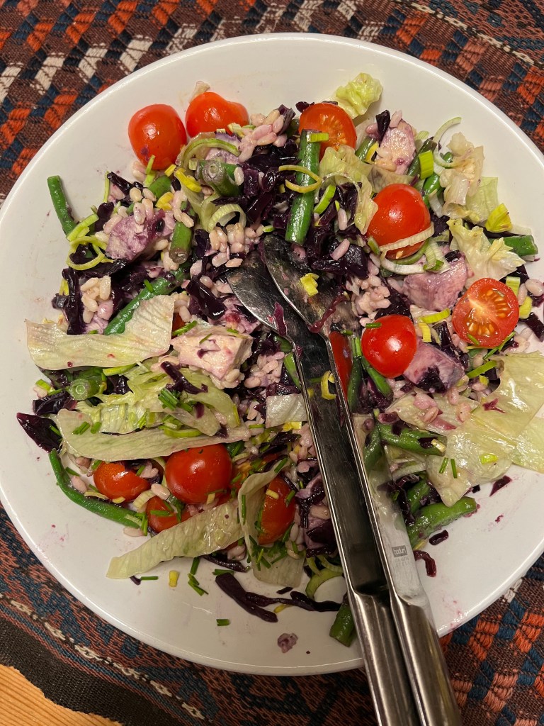 En färgglad sallad med rödkål, körsbärstomater, grönsallad, purjolök och blad av grönsaker, serverad i en vit skål med tång för servering.