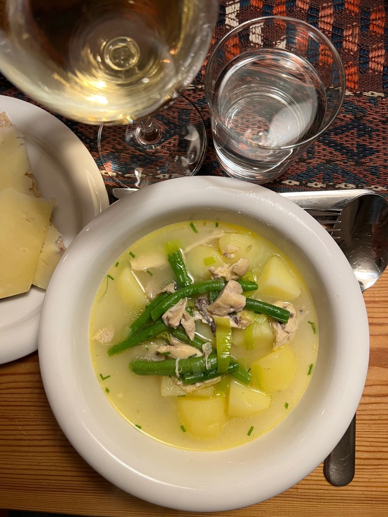 En skål med soppa innehållande potatis, gröna bönor och kyckling, serverad med ett glas vitt vin och vatten.