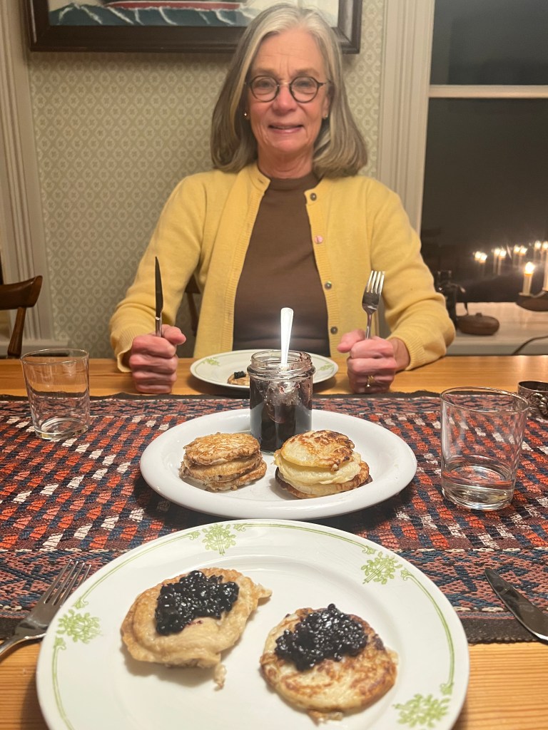 En kvinna sitter vid ett matbord med en tallrik innehållande plättar med sylt, med en glasburk med sylt framför sig. Bakgrunden visar en välbelyst miljö med fönster och levande ljus.